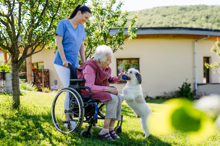 Smiling female caregiver helping an elderly woman in a wheelchair while they interact with a cute dog.
