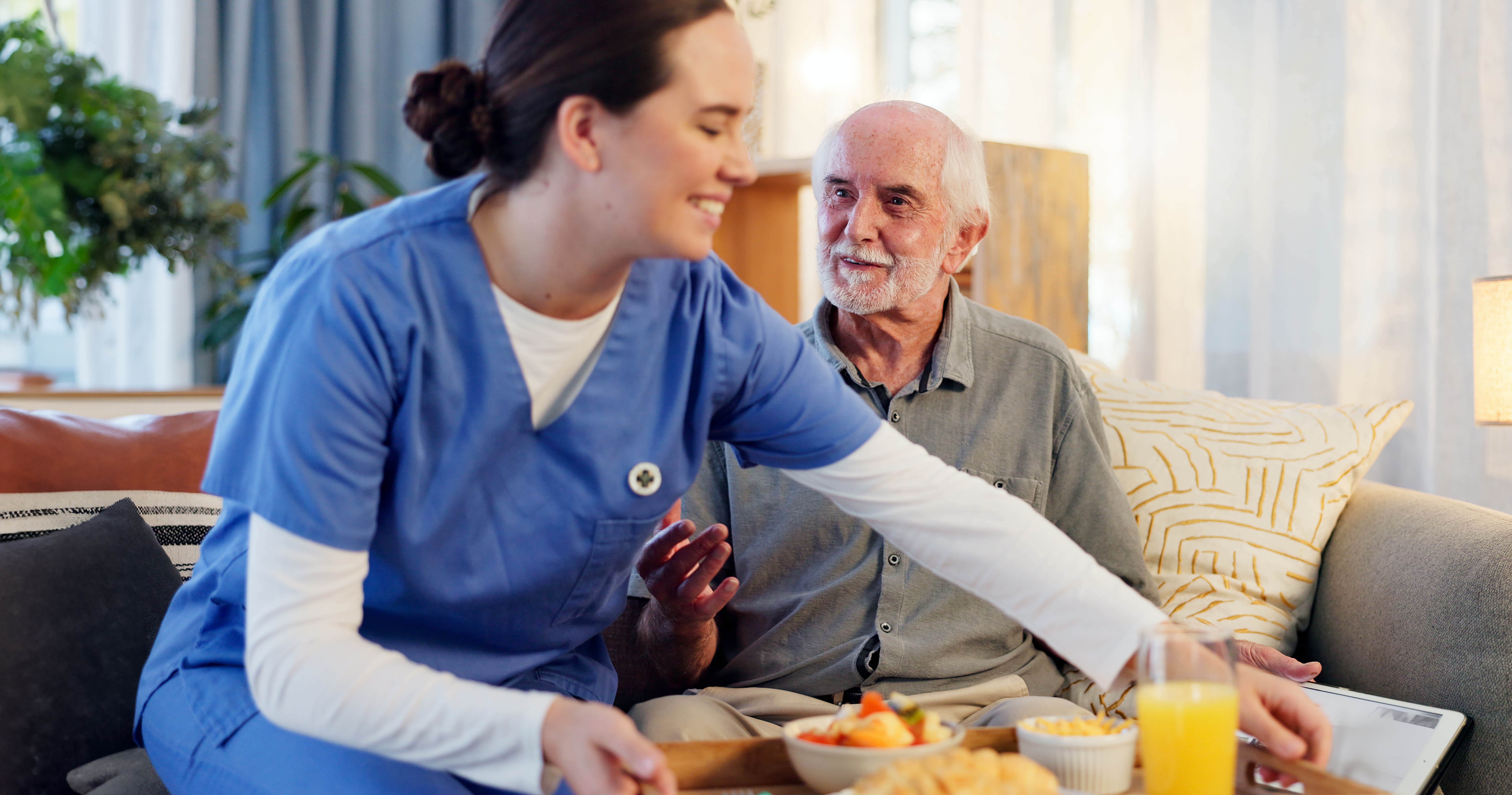 Smiling caregiver helping an elderly man move a food tray at home.
