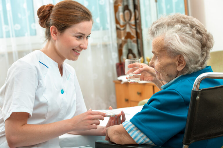 A smiling female caregiver with a warm expression, wearing light blue scrubs, is gently guiding an elderly woman with silver curly hair in a floral cardigan to take her medicine. The caregiver holds a small cup with pills and a glass of water, showing patience and care. The elderly woman looks up at the caregiver with a trusting expression. The background is a comfortably furnished living room with other residents and soft daylight.